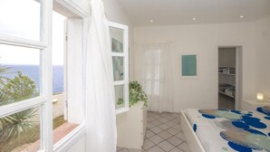 Une chambre avec vue sur la mer, un lit recouvert d’une couvre-lit à motifs bleus et blancs, une commode blanche, une plante et un placard avec une porte de couleur turquoise.