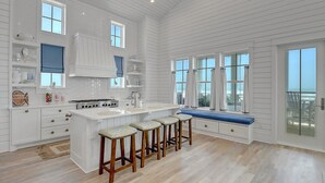 A spacious kitchen with a central island, wooden stools, and large windows offering views. - 8-bedroom home w/ Gulf-Front Views + Amenities (Port Aransas)