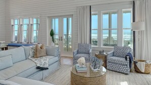 A spacious living room with a light blue sofa, patterned armchairs, and a coffee table. Large windows offer a view of the ocean. - 8-bedroom home w/ Gulf-Front Views + Amenities (Port Aransas)