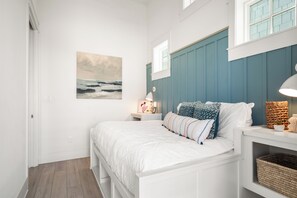 Un dormitorio con una cama blanca, un cuadro en la pared, una ventana y una mesita de noche con una lámpara.