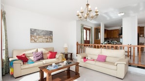 Una sala de estar con dos sofás color beige, una mesa de centro de madera y una araña. Se ve una cocina al fondo.