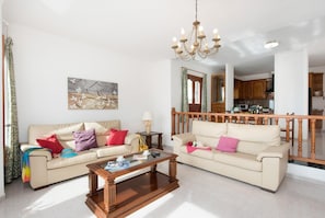 Una sala de estar con dos sofás color beige, una mesa de centro de madera y una araña. Se ve una cocina al fondo.