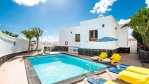 Una piscina con sillas de descanso y sombrillas, rodeada por un edificio blanco con un muro de piedra y palmeras.