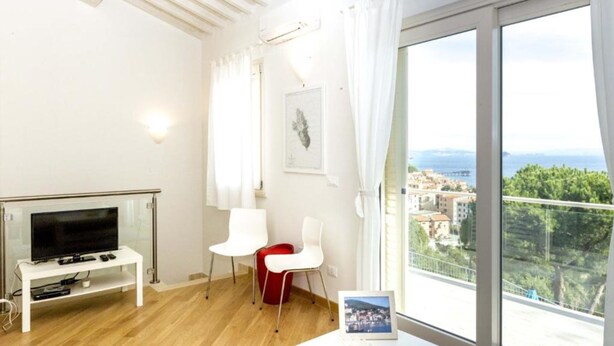 A modern room with a TV on a white stand, a small table with a picture frame, and a view of a coastal town through large windows.