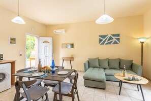 A well-lit living room with a sofa, a dining table with chairs, a washing machine, and a small table with a bottle and glasses. - Villa Russa Alexandros (Afrato)