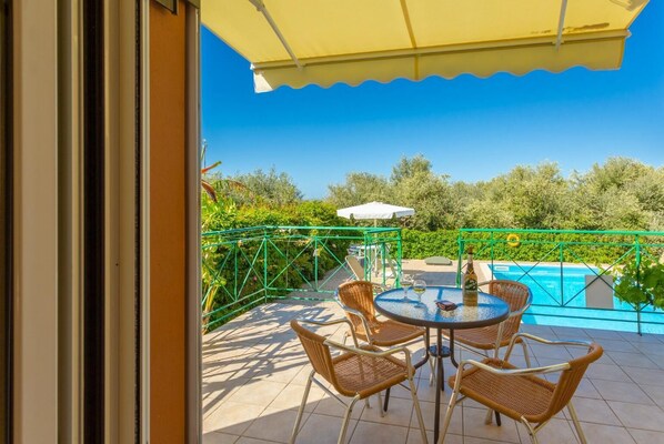 Une terrasse extérieure avec une table ronde et des chaises en osier, un parasol jaune et une vue sur une piscine.
