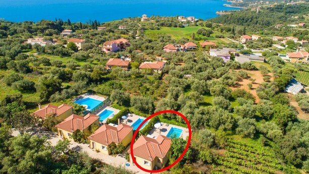 Oversigtsbillede af et kystnært boligområde med huse, oliventræer og et klart, blåt hav.
