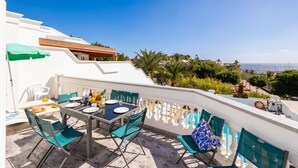 An outdoor dining area with a table set for two, surrounded by chairs, and a view of the sea and greenery in the background. - Villa Ramos Uno - Four Bedroom Villa, Sleeps 8 (Puerto Calero)