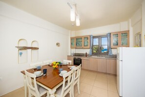 Eine Küche mit Esstisch, Stühlen, einer Obstschale, Schränken, einem Kühlschrank und einem Fenster mit Vorhängen.