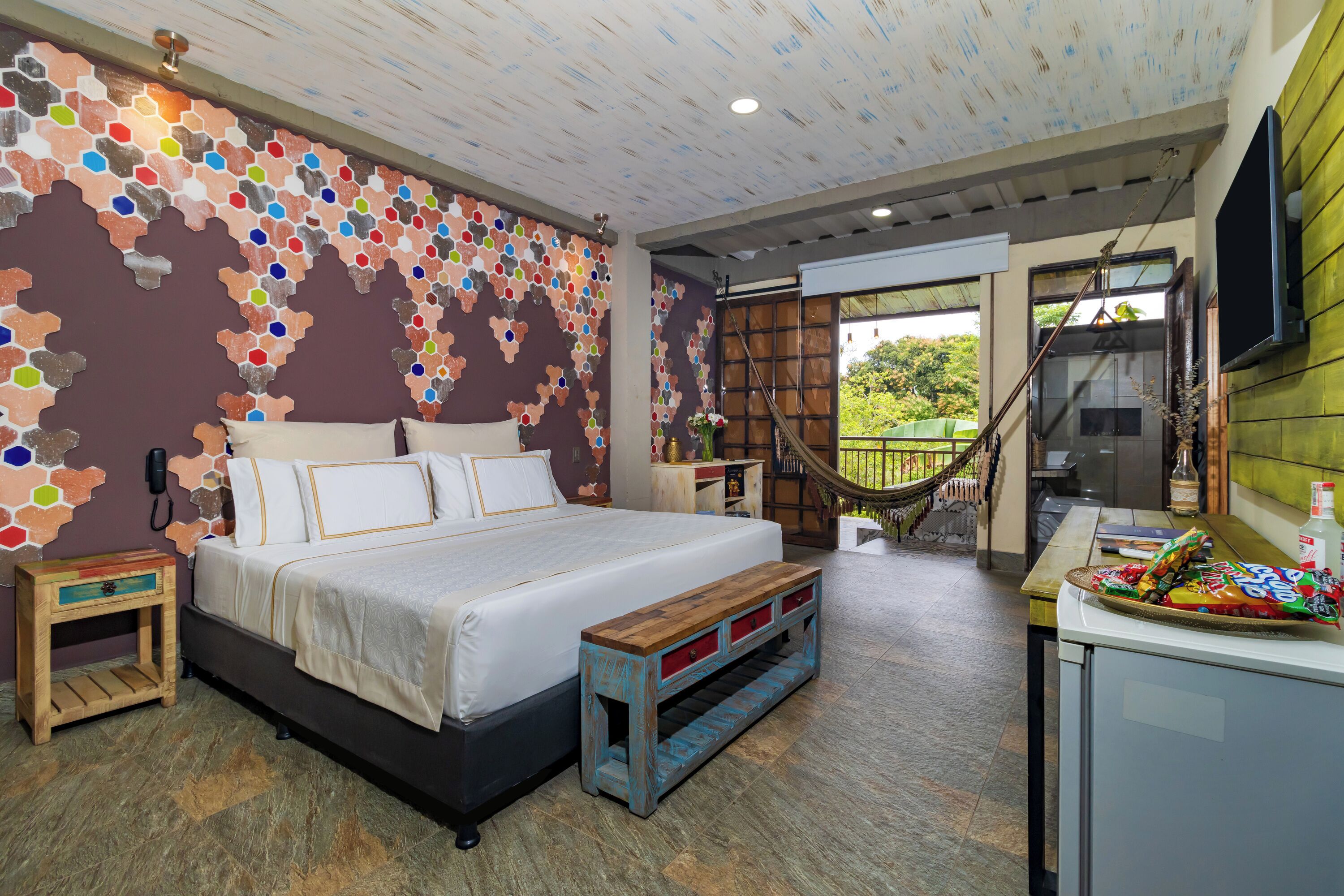 A bedroom with a bed, a wooden bench, a small table with a bottle, and a hammock.