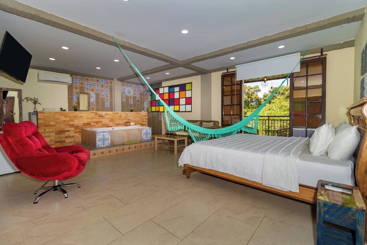 A room with a bed, a red chair, a hammock, a TV, and a bathroom with a tiled wall.