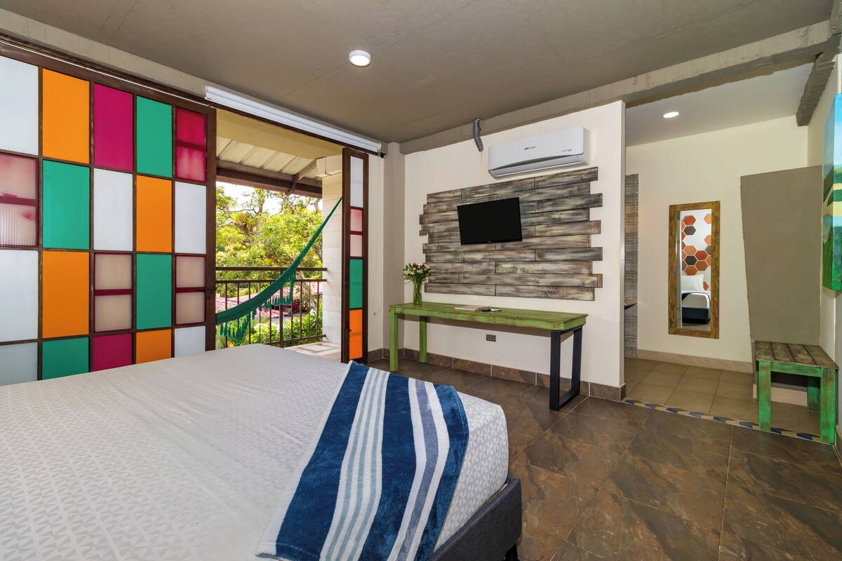 A bedroom with a bed, a TV mounted on a wall, a desk, and a window with a view of greenery.