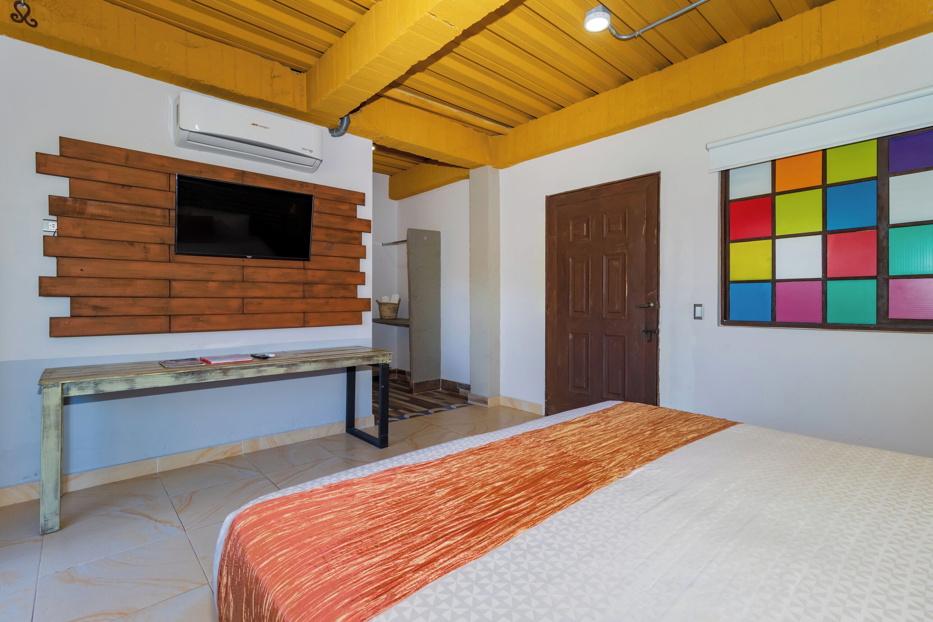 Un dormitorio con una cama, cabecera de madera, televisión montada en la pared y un ventanal colorido.