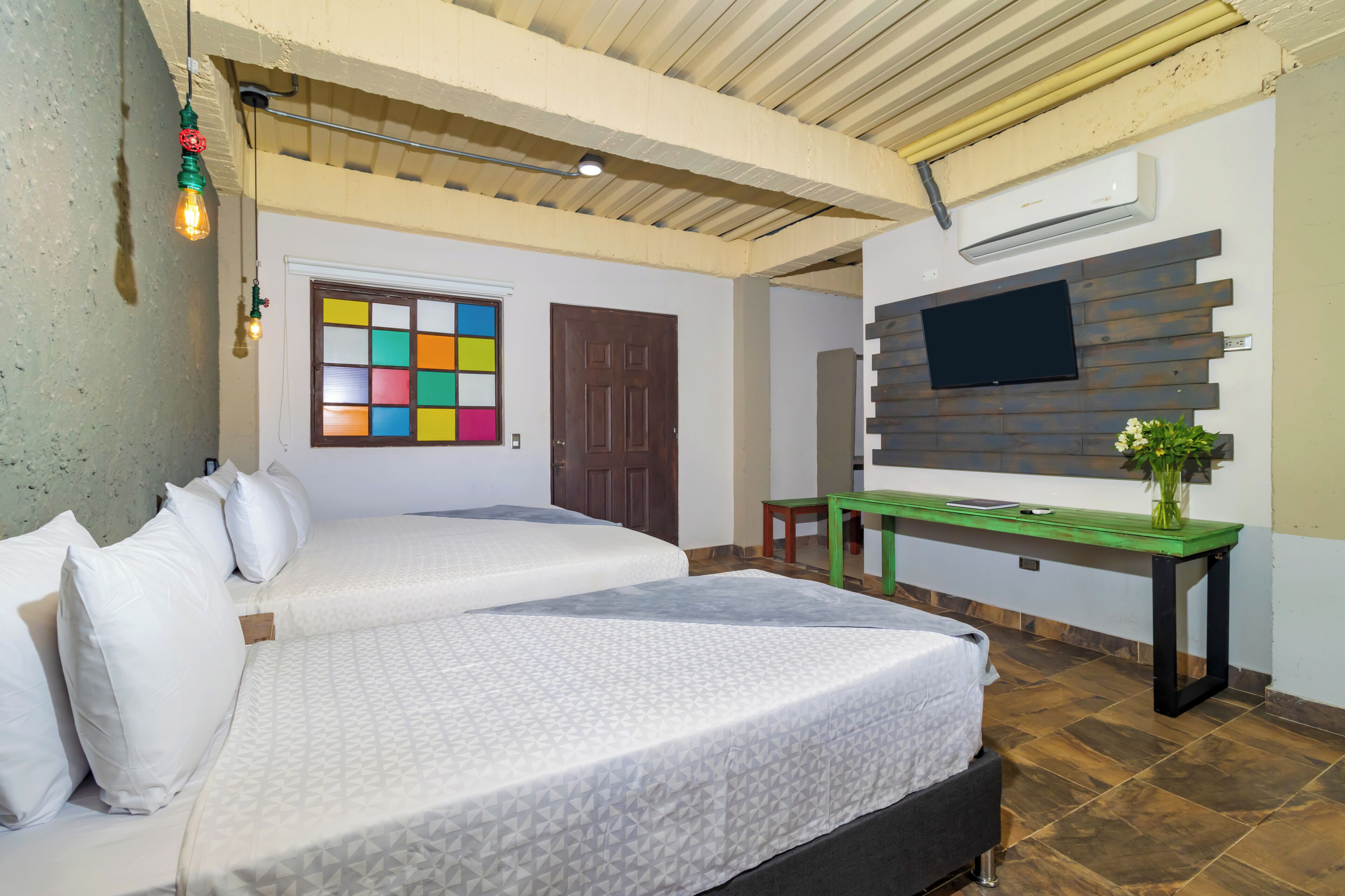 A hotel room with two beds, a desk, a TV, and a window with colorful blinds.