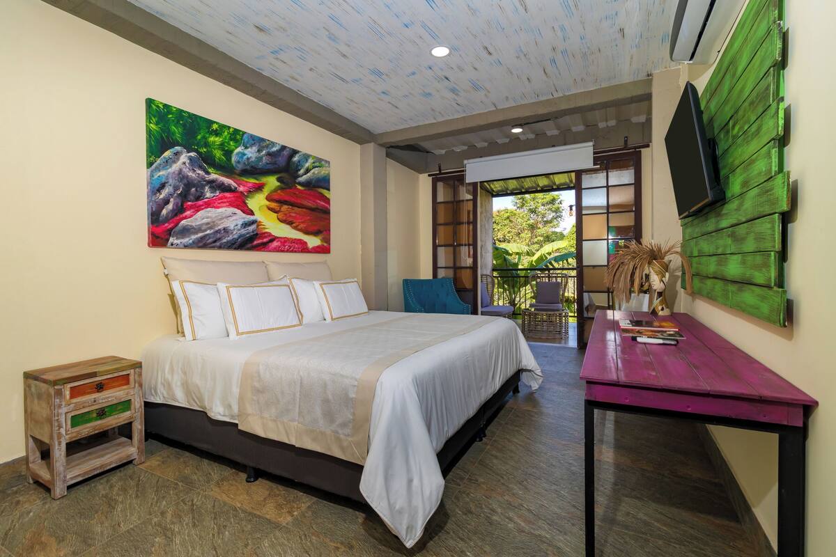 A bedroom with a large bed, a painting on the wall, a TV mounted on the wall, and a view of the outdoors.