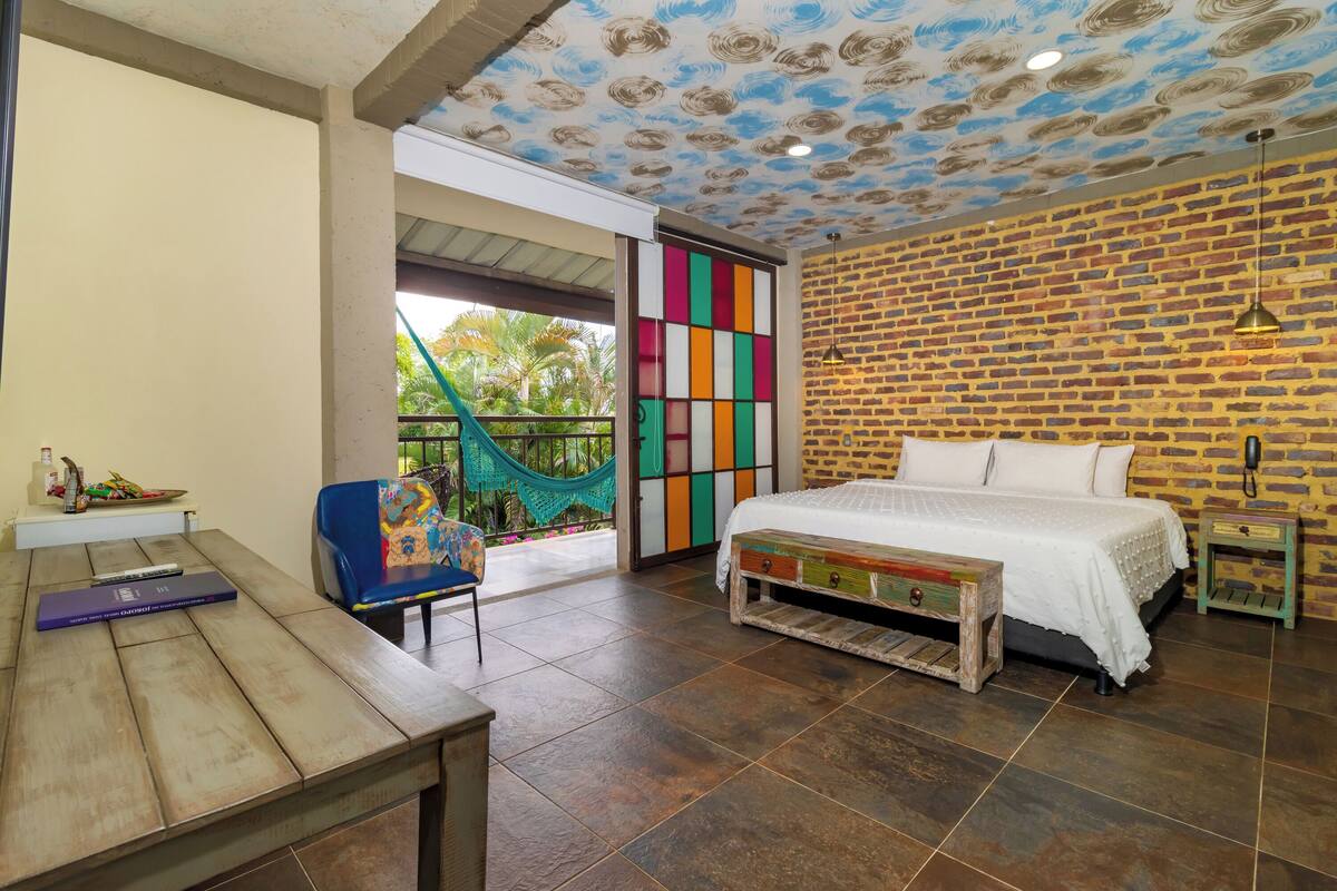 A bedroom with a brick wall, a bed, a wooden table, a chair, and a colorful partition.