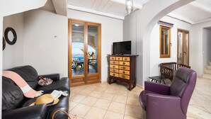 Un salón con un sofá de cuero negro, un sillón morado, un aparador de madera con cajones, un televisor sobre un mueble bajo y vistas a una zona de estar exterior a través de un ventanal.