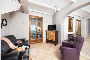 Ein Wohnzimmer mit einer schwarzen Ledersofa, einem violetten Sessel, einer Holzkabine mit Schubladen, einem Fernseher auf einem Schrank und Blick durch eine Glastür auf einen Außenbereich mit Sitzgelegenheiten.