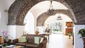 A living room with a brown leather sofa, a coffee table, and a fireplace. There are potted plants and a chandelier. - Elegant Sorrento Coast Villa with Pool and Tennis Court (Massa Lubrense)