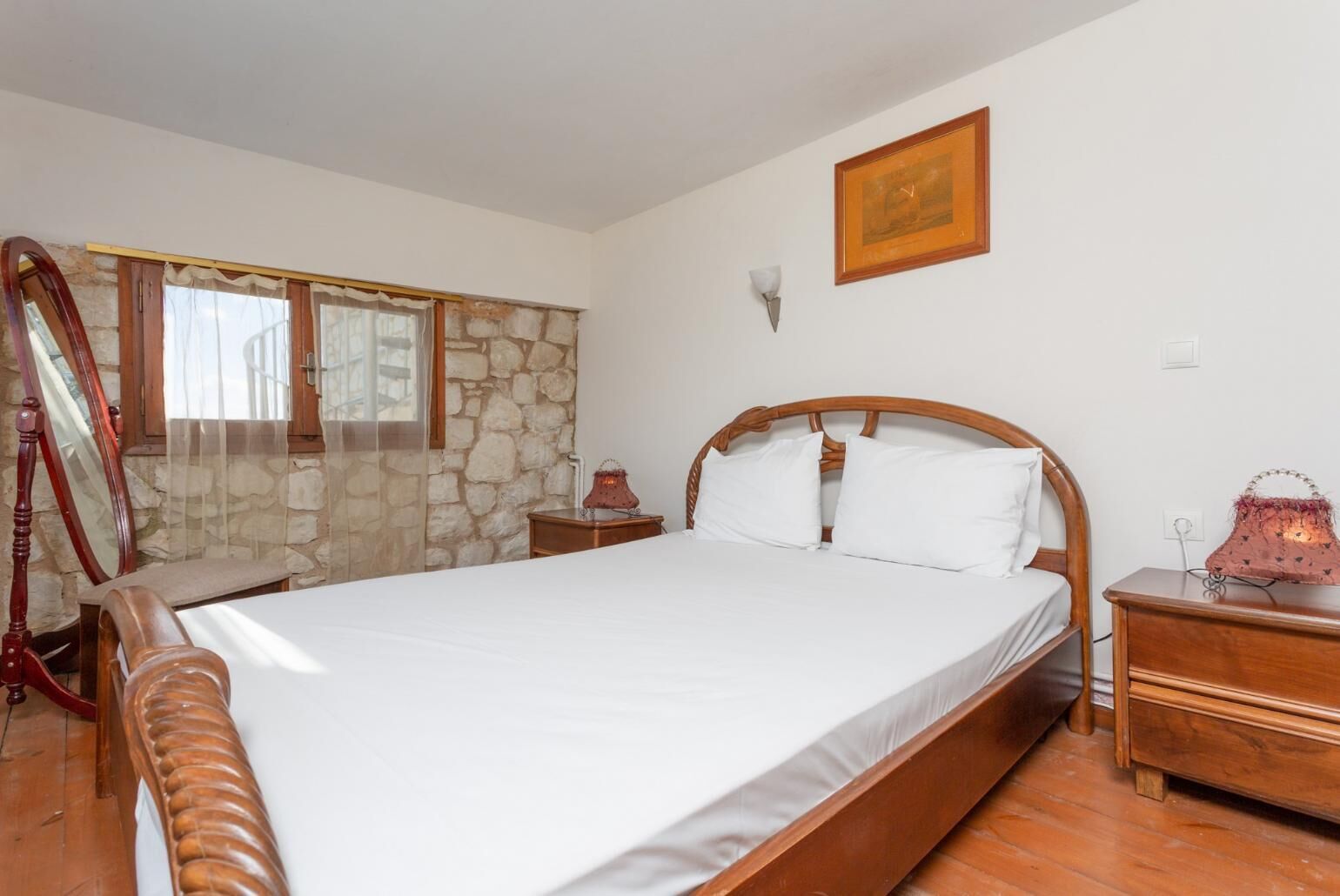 Un dormitorio con cama de madera, pared de piedra, ventana con cortinas, mesita de noche y una lámpara.