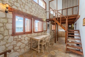 A stone-walled room with a wooden table and chairs, a staircase, and a view of the sea.