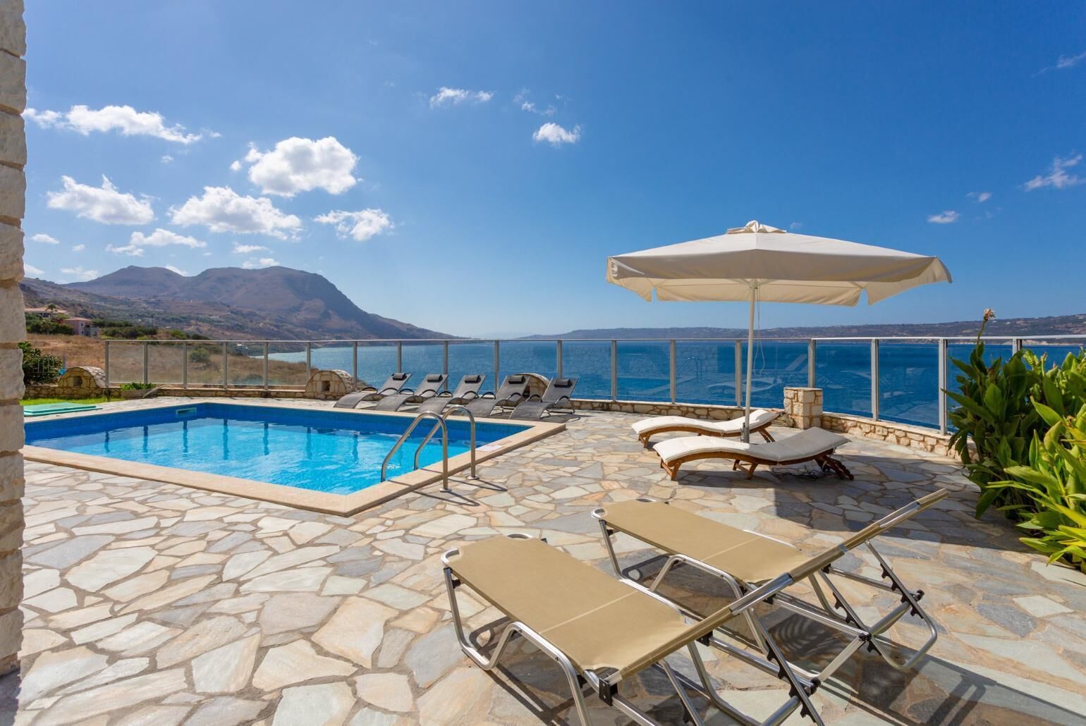 A pool area with lounge chairs and an umbrella, overlooking a scenic coastline with mountains.