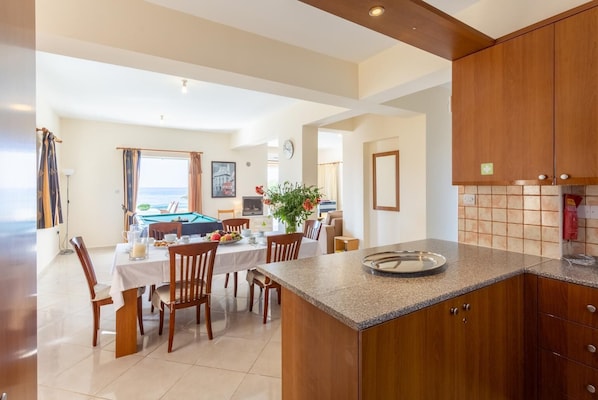 Une cuisine spacieuse dotée de meubles en bois et d’un plan de travail en granit. On y trouve un coin repas avec une table de billard et des chaises, ainsi qu’une vue sur la mer grâce à de grandes fenêtres.