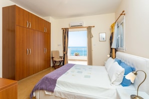 Une chambre avec un lit, une armoire en bois, une chaise, un balcon avec vue sur la mer et un appareil de climatisation.