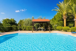 A swimming pool with a gazebo and lounge chairs, surrounded by greenery and palm trees.