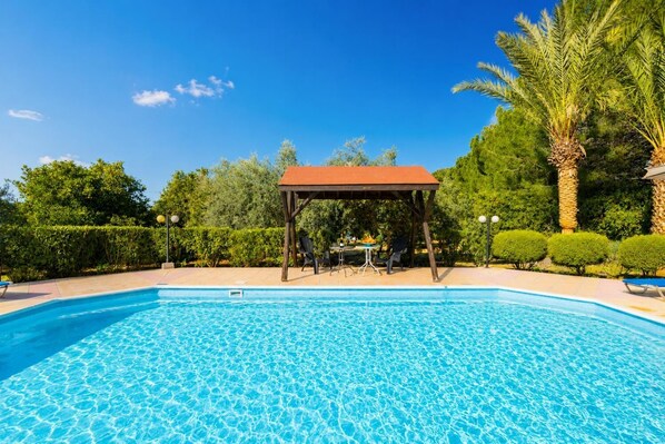 A swimming pool with a gazebo and lounge chairs, surrounded by greenery and palm trees.