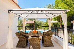 A covered outdoor seating area with wicker furniture, a pool in the background, and a view of greenery.