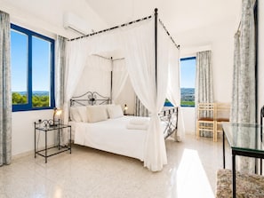 A four-poster bed with a canopy, a bedside table with a lamp, a chair, and a view of the landscape through the windows.