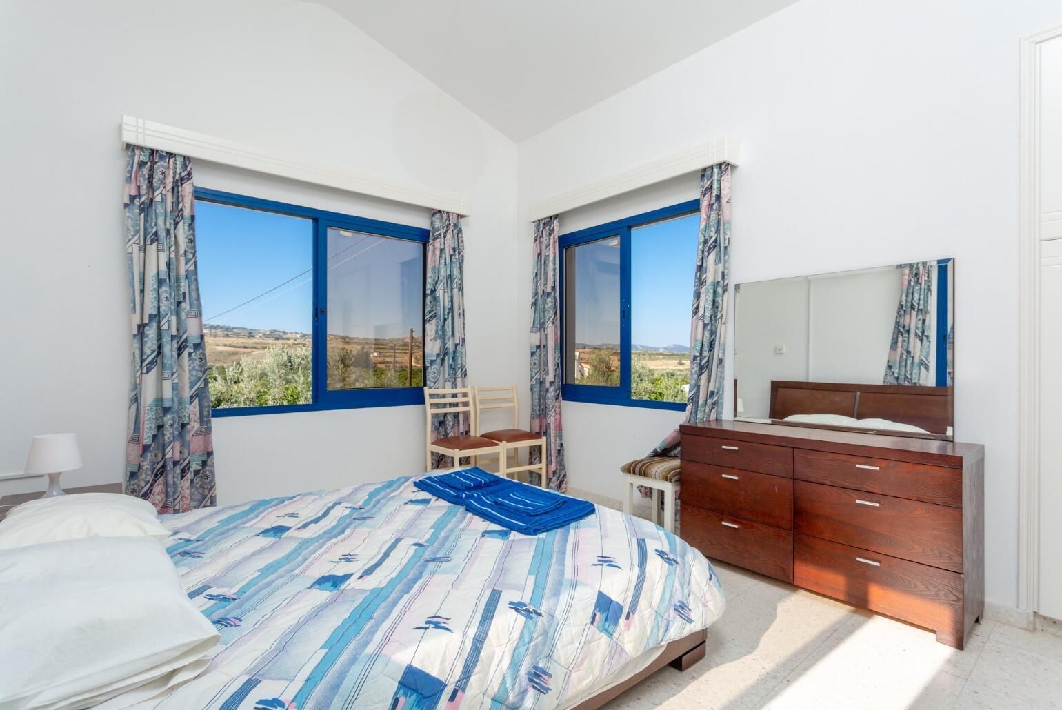 A bedroom with a bed, a chair, a dresser, and two windows with curtains.