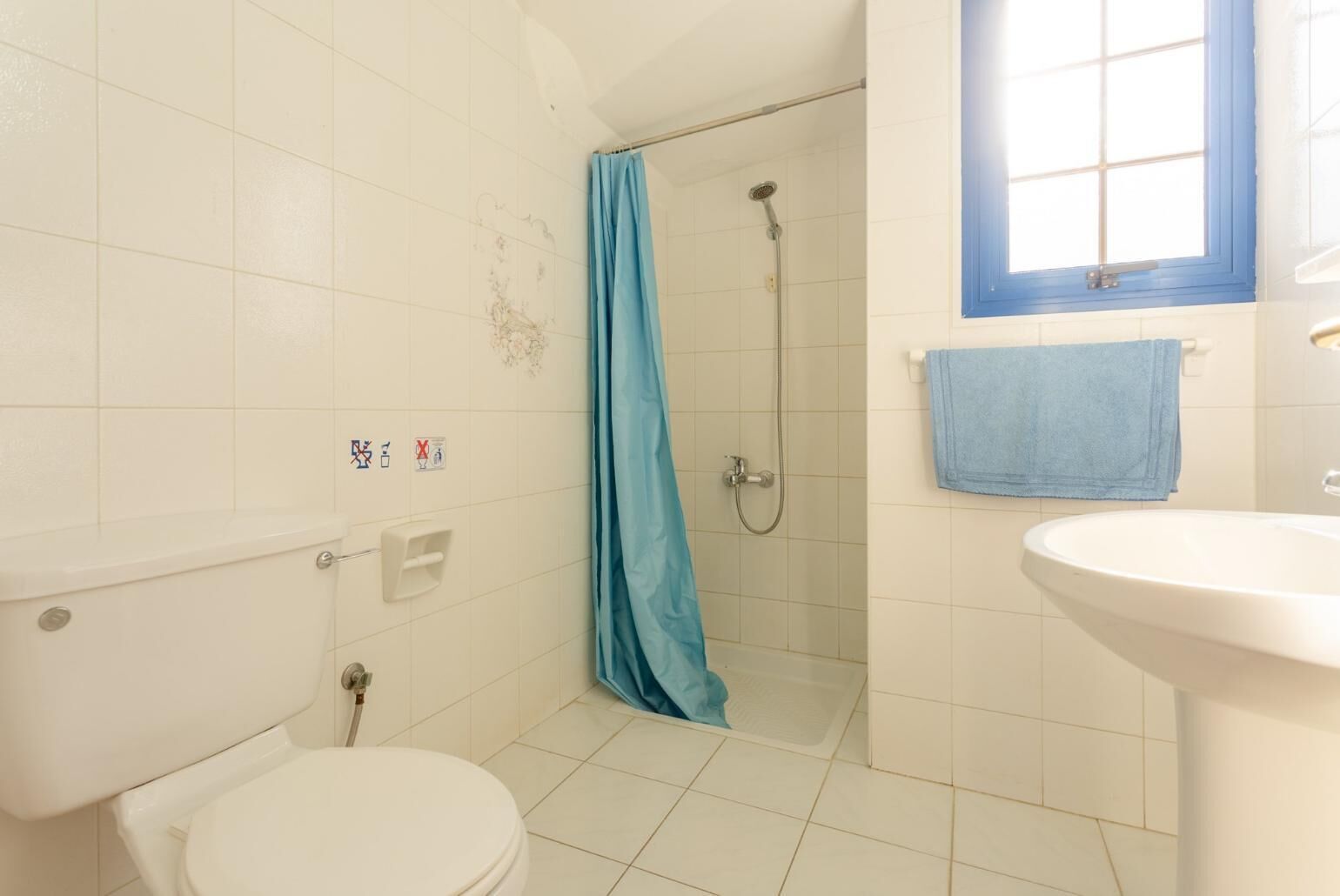 A bathroom with a toilet, a shower with a blue curtain, a sink, and a window with a blue frame.