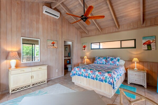 A bedroom with a wooden ceiling, a bed with a floral bedspread, a nightstand with a lamp, and a window with a view of greenery.
