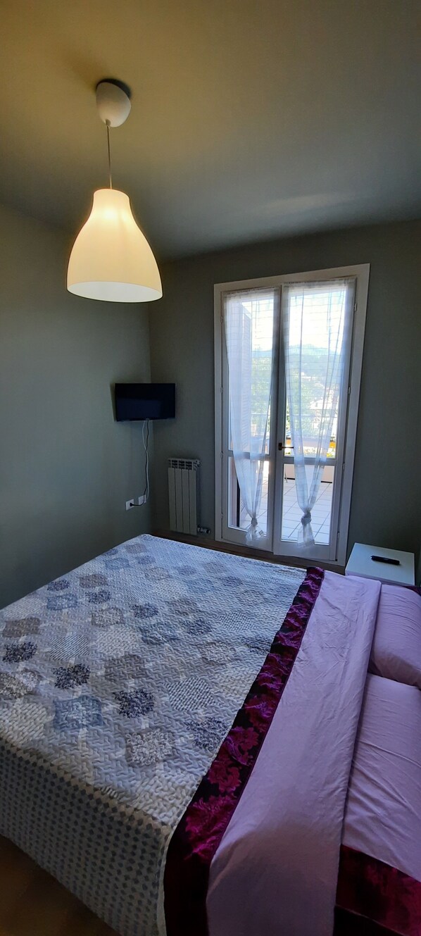 Un dormitorio con cama, luz en el techo, televisión y ventana con cortinas.