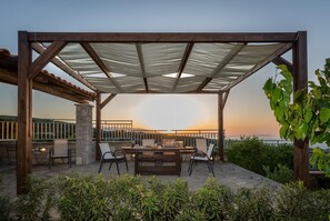 Un patio al aire libre con una pérgola de madera, pilares de piedra y vistas al atardecer sobre el mar.