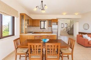 A modern kitchen with wooden cabinets, a central island, and a dining area with a table and chairs. - Villa Selini - Three Bedroom Villa, Sleeps 6 (Roussospiti)