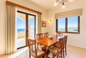 Un espace repas avec une table et des chaises en bois, une vue sur la mer grâce à de grandes fenêtres et un balcon agrémenté de rideaux.
