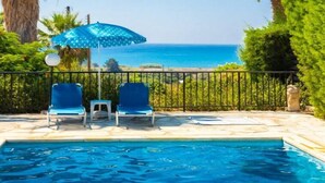 Una piscina con dos sillas reclinables azules y una sombrilla azul, rodeada de palmeras y vistas al mar.