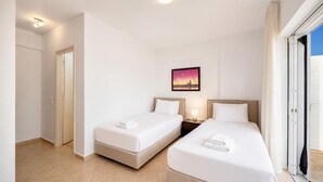 Een hotelkamer met twee bedden, een nachtkastje met een lamp, een schilderij aan de muur en een deur die naar een andere kamer leidt.