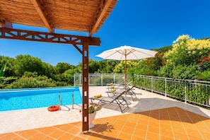 A pool area with a pergola, outdoor seating, and a view of the surrounding greenery.