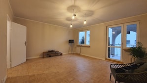 A spacious room with wooden flooring, a bench, a standing lamp, and a large window with a view of buildings. - matejki apartament (Poznan)