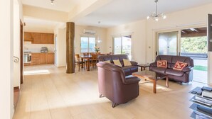 A spacious living area with a sofa set, a coffee table, and a TV stand. There is a dining area with a table and chairs, and a kitchen in the background.