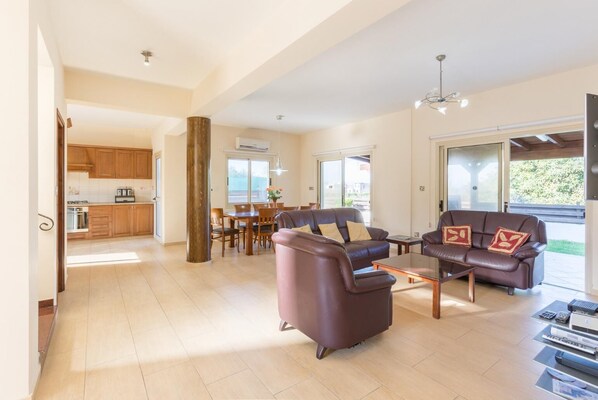Un espacio habitable amplio con un conjunto de sofás, una mesa de centro y un mueble para la televisión. Se observa una zona de comedor con una mesa y sillas, y una cocina al fondo.