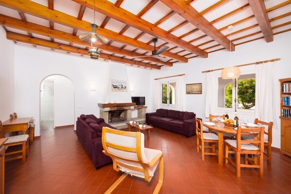 Un vaste espace de vie doté d’une cheminée, de poutres apparentes en bois au plafond, d’une table à manger avec des chaises et d’un canapé.