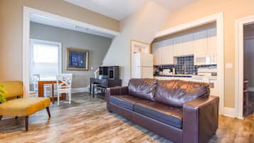 A living room with a brown leather sofa, a dining area with a wooden table and chairs, and a kitchen with white cabinets and a black backsplash.