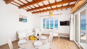Un coin repas avec une table dressée pour quatre personnes, un panier de fruits et une bouteille de vin. On y trouve des chaises blanches, un plafond en bois et une vue sur la mer par de grandes fenêtres.