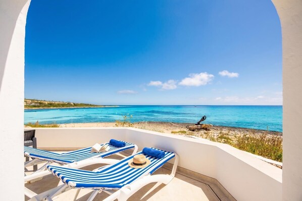 Un balcon avec deux fauteuils de jardin à rayures, un chapeau et une vue sur la mer et le littoral au loin.