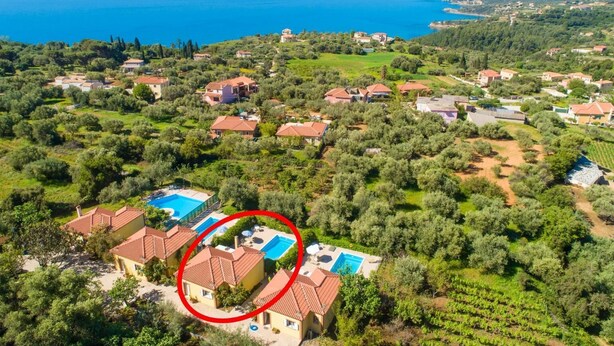 Vista aérea de una zona residencial costera con casas, olivares y un mar azul cristalino.